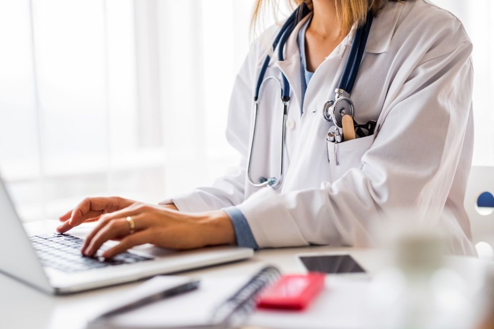 Doctor typing on a laptop with a stethoscope, focused on patient care or medical research in a bright office.