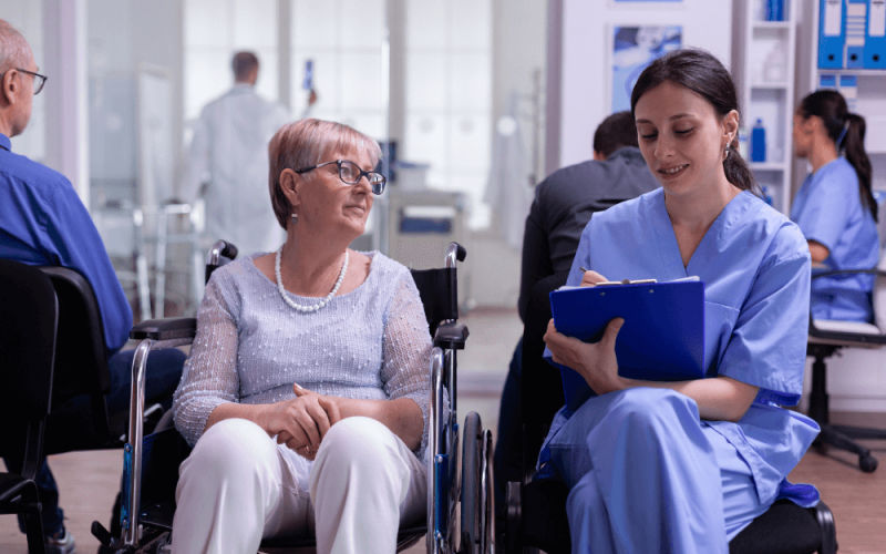 A woman in a wheelchair and a nurse in scrubs engage in conversation; the nurse takes notes on a clipboard in a bright, clinical environment with other patients and healthcare professionals nearby.