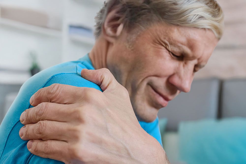 Man in blue shirt holding shoulder in pain, indicating discomfort or injury in home setting.