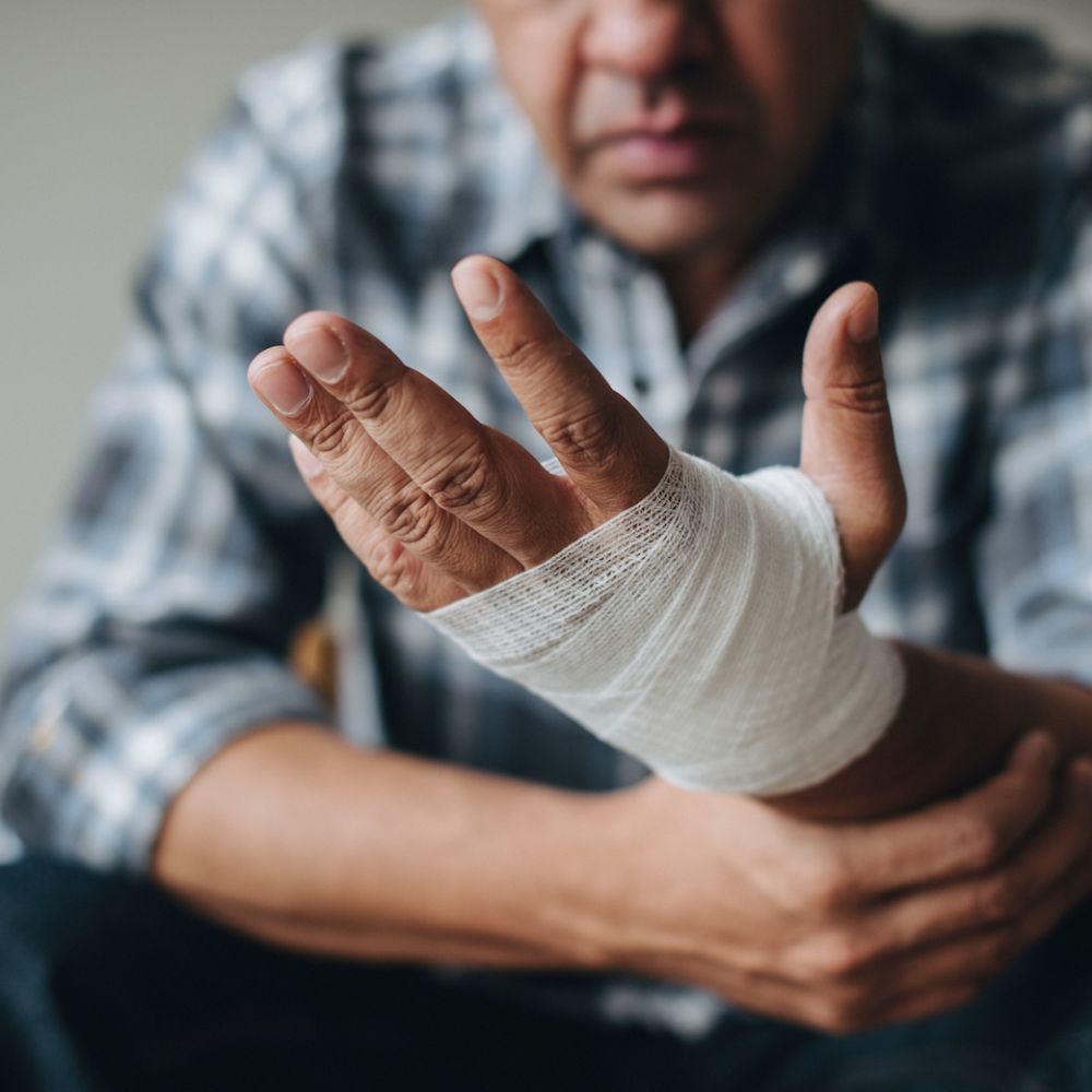 Man with bandaged hand sitting in plaid shirt, looking at injured wrist indoors.