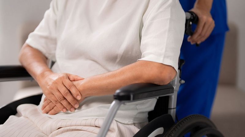 A person sits in a wheelchair with hands folded, wearing a white shirt. Another person in blue clothing stands behind, holding the wheelchair in a brightly lit room.