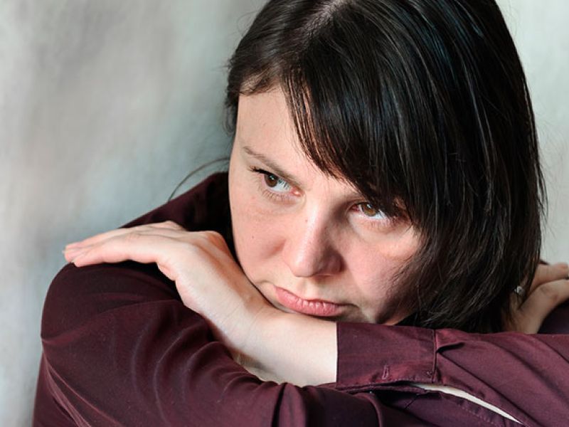 Woman deep in thought, resting head on arms, wearing a maroon shirt. Contemplative expression, light background.