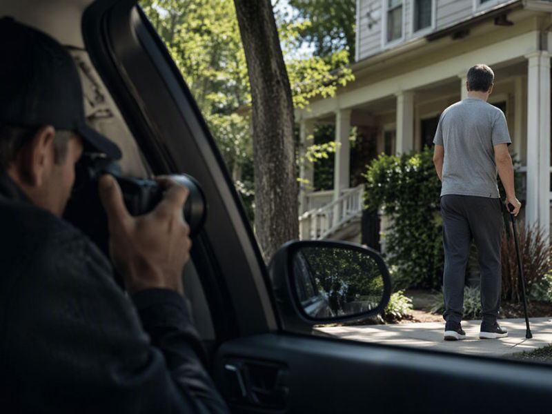 A person with a walking cane approaches a house; another person photographs them from a car.