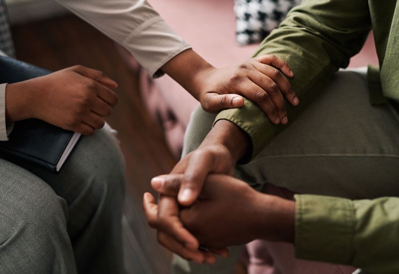 Two people in a comforting gesture with hands touching during a supportive conversation in a cozy setting.