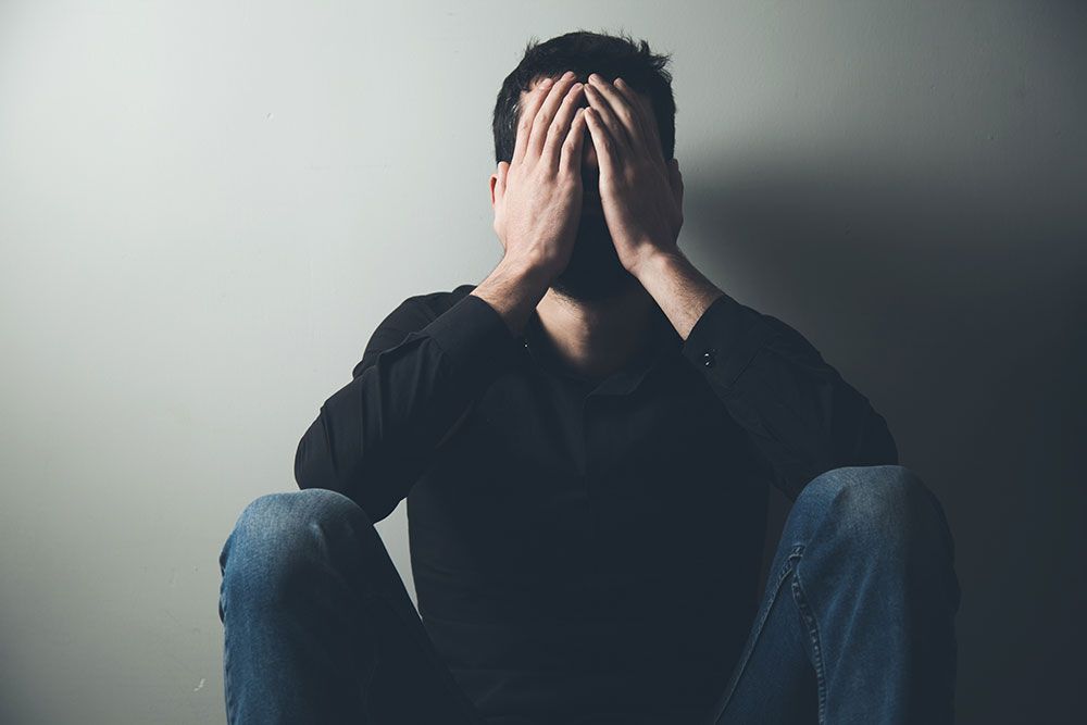 Man sitting against a wall, covering face with hands, conveying stress or frustration.