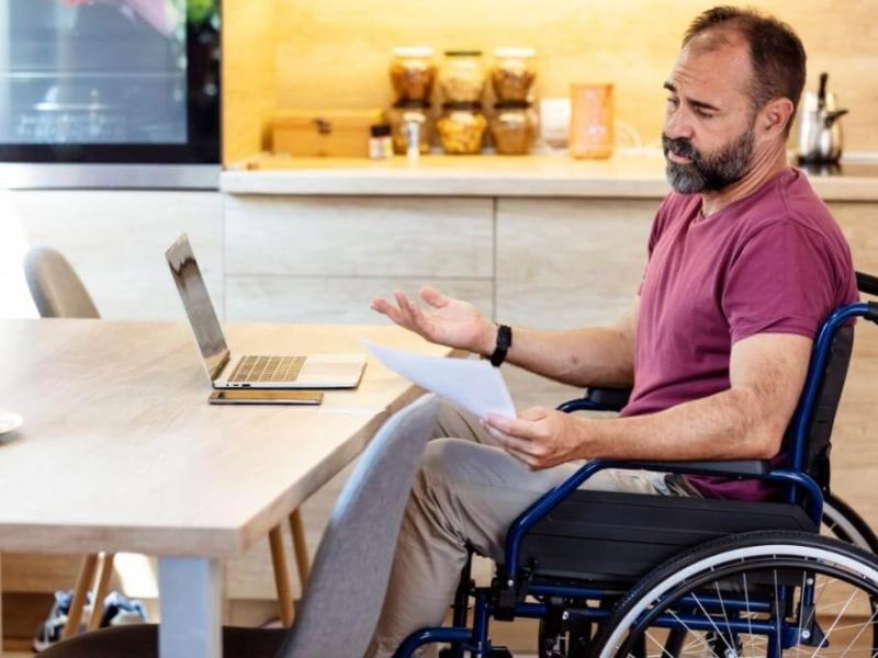 Man in a wheelchair sits at a wooden kitchen table with a laptop and papers, gesturing as he explains something.