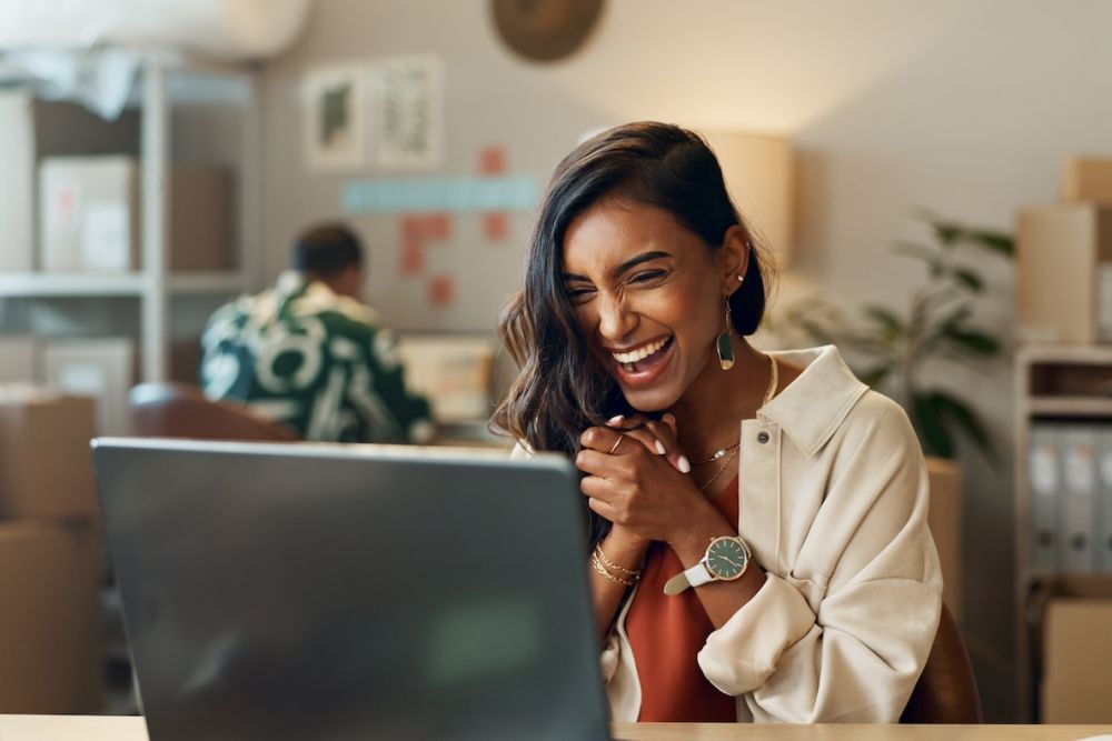 Smiling woman celebrating while looking at a laptop in a modern office workspace.