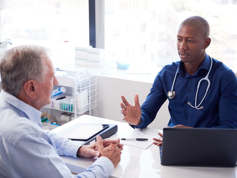 Doctor consulting with an elderly patient in an office, discussing health concerns. Stethoscope and laptop on desk.