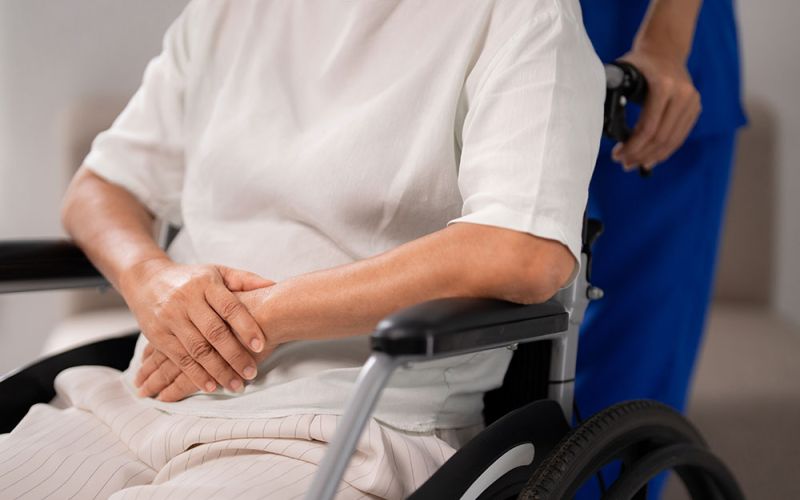 A person sits in a wheelchair with hands folded, wearing a white shirt. Another person in blue clothing stands behind, holding the wheelchair in a brightly lit room.