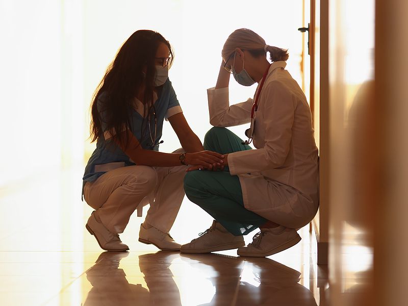 Two healthcare workers in masks sit on a hallway floor, offering support and comfort to each other in a hospital.