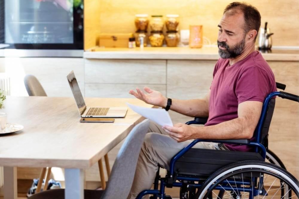 Man in a wheelchair sits at a wooden kitchen table with a laptop and papers, gesturing as he explains something.