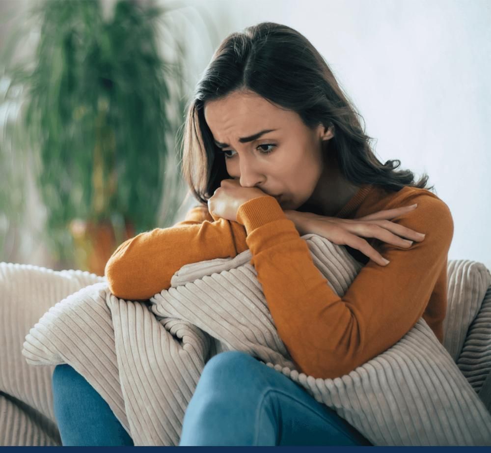 Woman in orange sweater looks thoughtful, holding pillow on sofa.