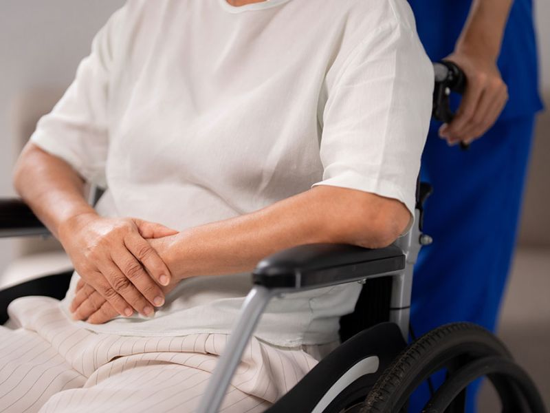 A person sits in a wheelchair with hands folded, wearing a white shirt. Another person in blue clothing stands behind, holding the wheelchair in a brightly lit room.