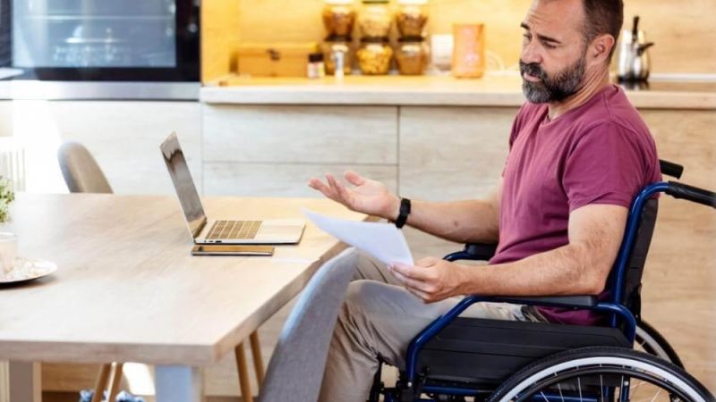 Man in a wheelchair sits at a wooden kitchen table with a laptop and papers, gesturing as he explains something.