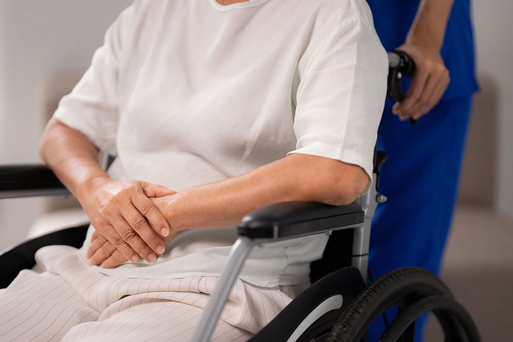 A person sits in a wheelchair with hands folded, wearing a white shirt. Another person in blue clothing stands behind, holding the wheelchair in a brightly lit room.