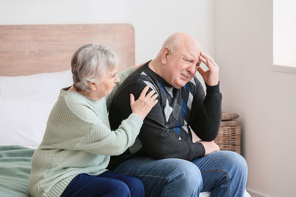 Elderly woman comforting a worried man sitting on a bed in a bright bedroom setting.
