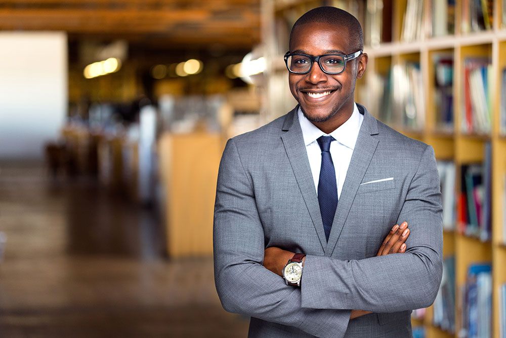 Confident professional in a grey suit standing in a library with arms crossed, smiling.