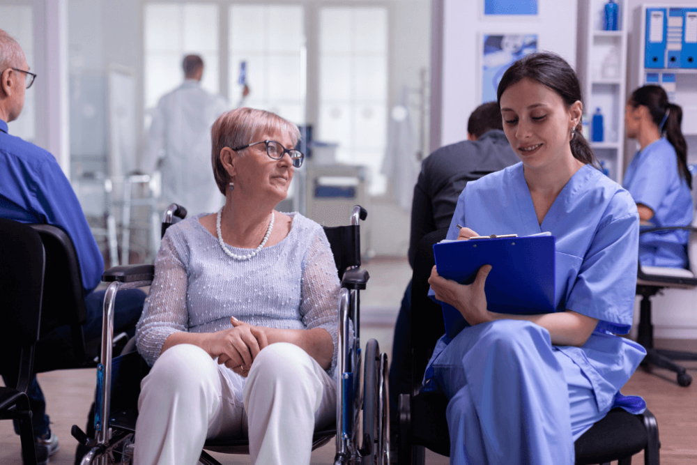 A woman in a wheelchair and a nurse in scrubs engage in conversation; the nurse takes notes on a clipboard in a bright, clinical environment with other patients and healthcare professionals nearby.