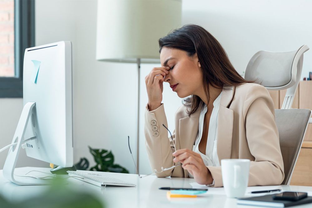 Businesswoman in an office feeling stressed, seated at a desk with a computer and holding glasses to her forehead.