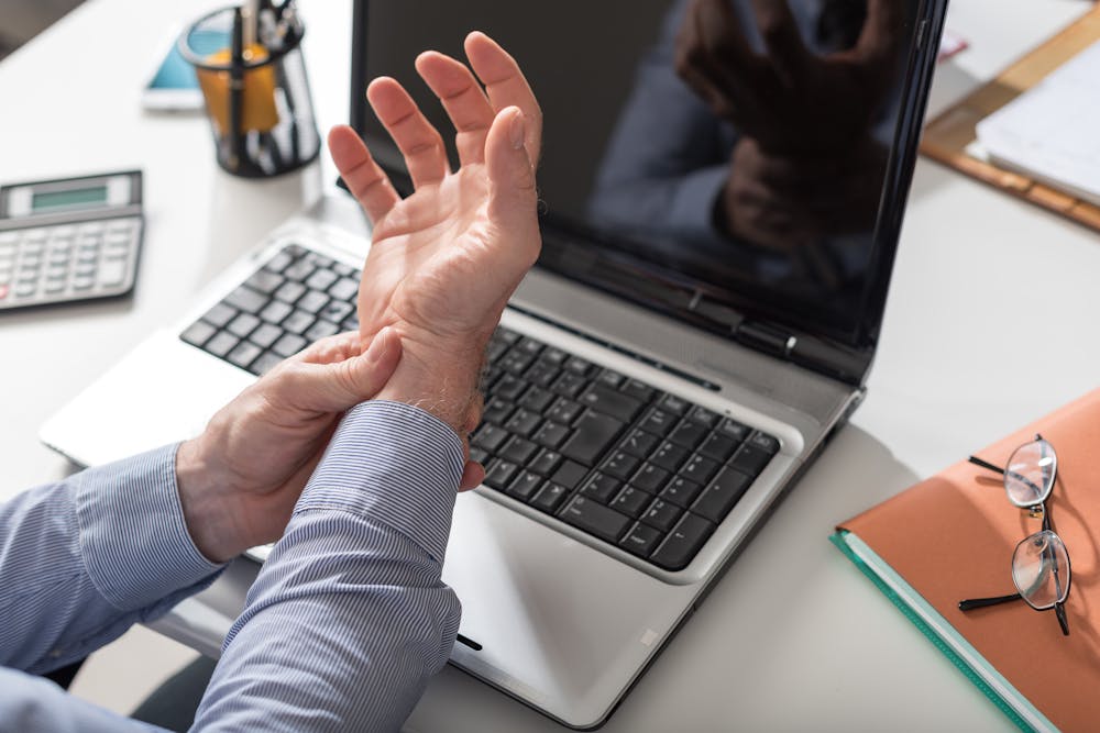 Do You Need a Carpal Tunnel Disability Claims Lawyer - Law Office of Justin Frankel A person grasps their wrist, suggesting discomfort, in front of an open laptop, with office supplies indicating a work environment.