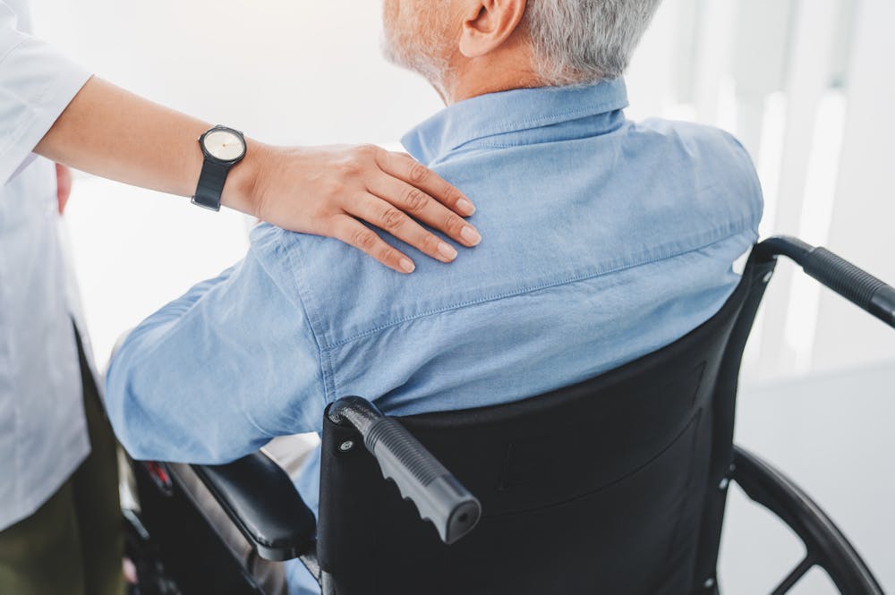 An elderly man sits in a wheelchair, while a caregiver gently places a hand on his shoulder, conveying support in a bright, modern environment with soft lighting.