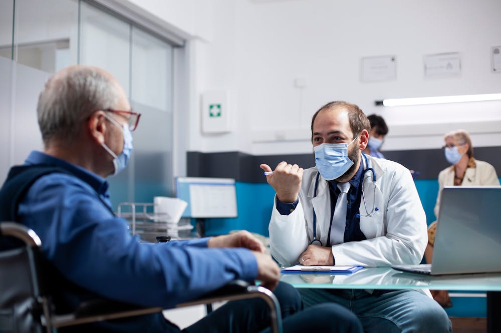 A male doctor in a white coat and mask sits at a desk, engaging in conversation with an elderly patient in a wheelchair. The scene takes place in a modern medical office.