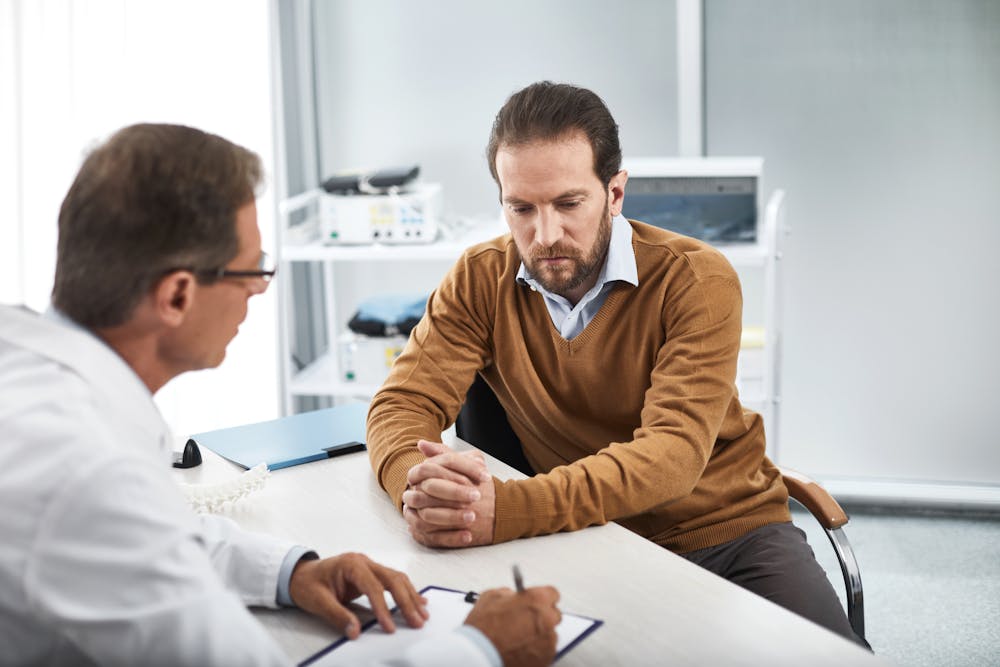 A man consults with a doctor across a desk in a well-lit, clinical room; medical equipment is visible in the background. Both individuals appear engaged in conversation.