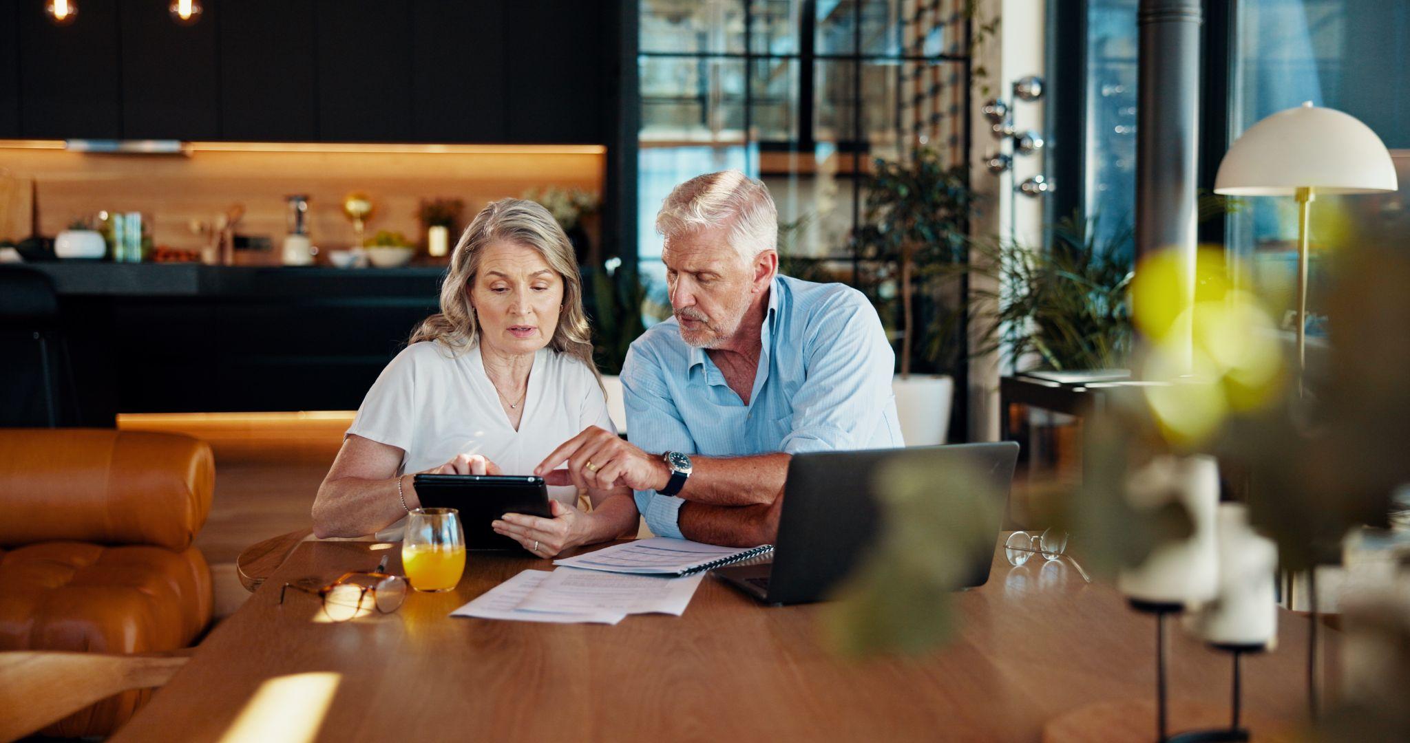 Older couple reviewing documents and using a tablet together at a dining table in a modern home.